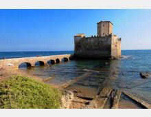 Castello costiero in pietra su un isolotto, collegato alla riva da un ponte basso ad arcate sopra il mare poco profondo. In primo piano si vedono una piccola spiaggia rocciosa con resti di strutture rettangolari e vegetazione, sullo sfondo il mare aperto e il cielo sereno.