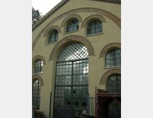 Facciata di un edificio industriale con grandi finestre ad arco incorniciate in mattoni e una porta centrale vetrata a griglia sotto un arco maggiore. Intonaco beige, struttura simmetrica con più aperture ad arco e recinzione metallica in primo piano.