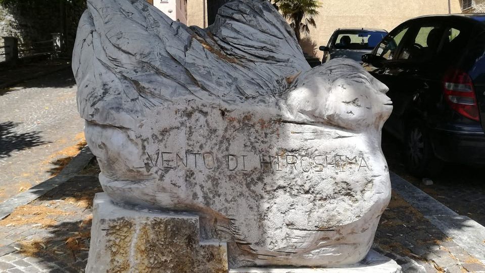 Scultura in pietra chiara a forma di uccello stilizzato con ali spiegate su basamento, collocata in una piccola area pavimentata all’aperto. Sullo sfondo si vedono un muro e edifici, alberi che fanno ombra e alcune auto parcheggiate.