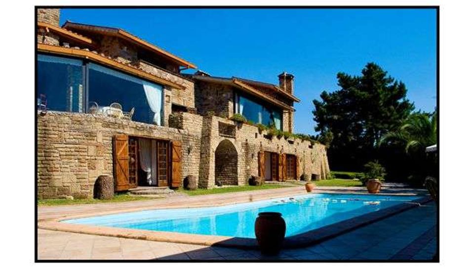 Villa in pietra a più livelli con grandi vetrate e porte in legno, affacciata su una lunga piscina rettangolare. Ampio patio pavimentato con vasi e giardino con alberi sullo sfondo sotto cielo sereno.