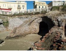 Rovine archeologiche in mattoni con un grande arco che conduce a un passaggio sotterraneo, circondate da muri in pietra. Sullo sfondo si vedono edifici moderni e una ringhiera bianca lungo il bordo superiore del sito.