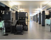 Ampio spazio espositivo interno con pavimento piastrellato e soffitto basso illuminato, scandito da pilastri scuri. Lungo il corridoio sono esposti grandi macchinari industriali e tubazioni metalliche.