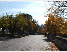 Strada in pavé leggermente in discesa, fiancheggiata da alberi con foglie autunnali e da un muretto in pietra sul lato destro. Sullo sfondo si vedono lampioni e edifici tra la vegetazione sotto un cielo limpido.