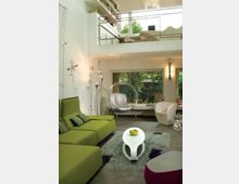 Soggiorno moderno a doppia altezza con soppalco e ringhiera in metallo, pareti bianche e pavimento in piastrelle scure. Divano verde, tavolino bianco su tappeto, poltrone di design e ampie finestre che affacciano su un giardino esterno.