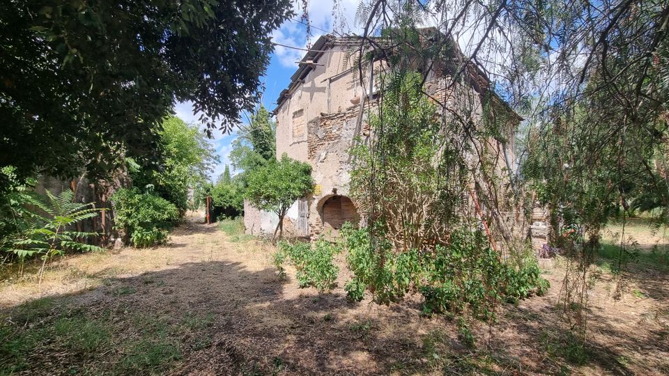 Edificio rurale in muratura parzialmente in rovina, con intonaco scrostato e archi al piano terra, circondato da vegetazione fitta. Un sentiero sterrato attraversa l’area ombreggiata da alberi e cespugli sotto un cielo parzialmente nuvoloso.