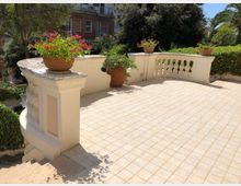 Terrazza esterna pavimentata con piastrelle chiare e parapetto curvo in muratura con balaustra; sui muretti sono appoggiati vasi in terracotta con fiori e piante. Intorno si vedono siepi e alberi, con un edificio residenziale sullo sfondo.