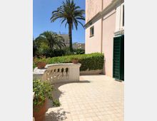 Terrazza esterna pavimentata con balaustra in muratura e vasi di fiori, affiancata da una facciata color rosa con porta a grata verde. Sullo sfondo si vedono siepi e vegetazione con alte palme e altri edifici sotto cielo sereno.