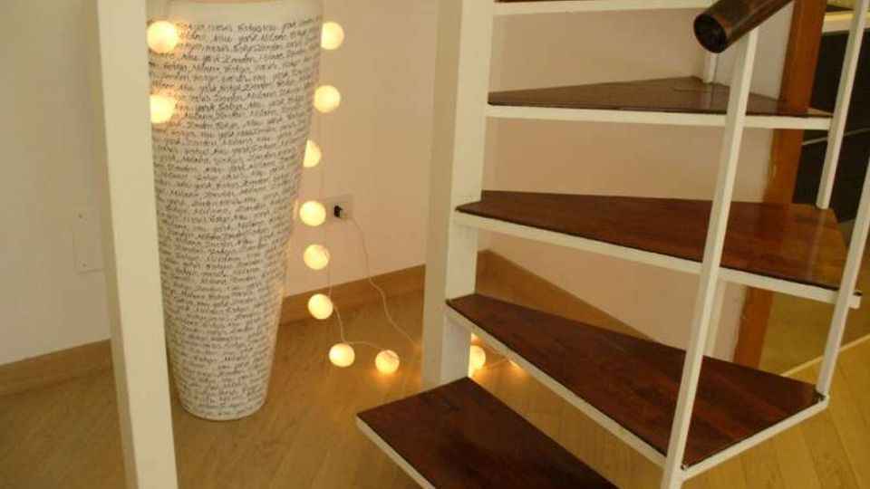 Interno domestico con scala a chiocciola in metallo bianco e gradini in legno scuro su pavimento in parquet. Accanto alla scala c’è una lampada cilindrica alta con scritte e una catena di luci a sfera appoggiata a terra.