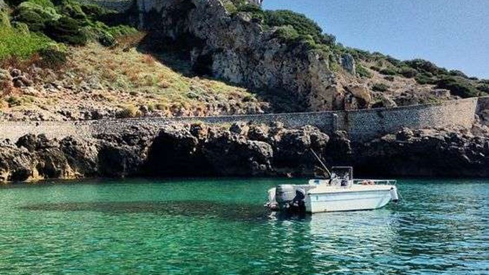 Baia costiera con acqua turchese e scogliere rocciose, con un piccolo motoscafo ancorato in primo piano. In alto sul promontorio si vede una torre circolare in pietra affiancata da un edificio basso, circondata da vegetazione mediterranea e un muro di contenimento lungo la costa.