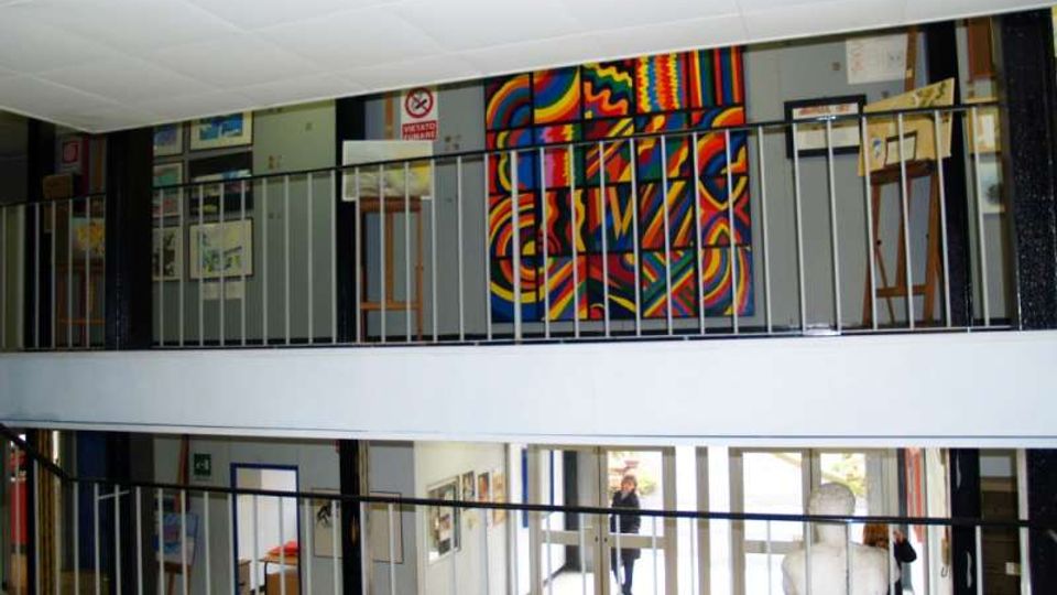 Interno di un edificio con atrio su due livelli, caratterizzato da passerelle con ringhiere metalliche e pareti espositive con quadri e cartelloni. Sul piano superiore è visibile un grande pannello artistico astratto e colorato, con finestre e porte vetrate sullo sfondo.