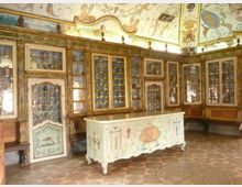Interno di una sala storica con pareti rivestite da grandi vetrine e armadi dorati pieni di piccoli oggetti esposti. Al centro c’è un banco decorato, mentre il soffitto è affrescato con motivi ornamentali e figure.