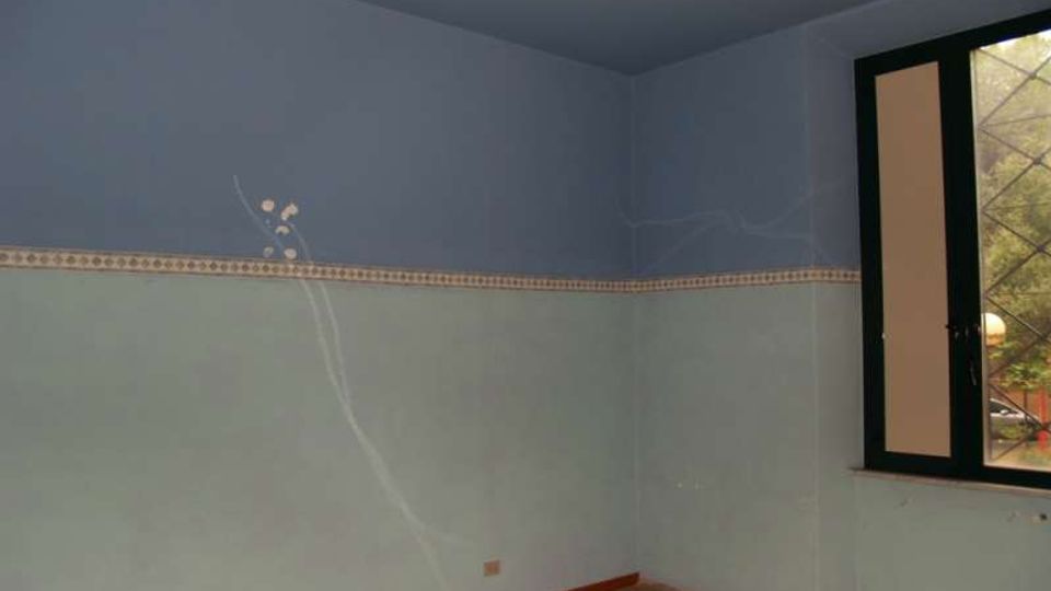 Interno di una stanza vuota con pareti bicolori azzurre, fascia decorativa orizzontale e pavimento grezzo. A destra c’è una finestra con inferriata e telaio scuro, con vista su alberi all’esterno.