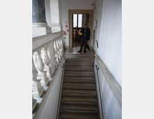 Scala interna in pietra con gradini larghi, corrimano in metallo e balaustra in marmo decorata. In cima alle scale si vede un pianerottolo con pavimento a mosaico e una porta con cornice in pietra che conduce a una stanza illuminata da una finestra.