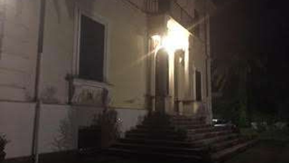 Facciata esterna di un edificio a più piani con intonaco chiaro, ingresso illuminato da una lampada e una scalinata in pietra che conduce a un portico con archi. Davanti si vede un piccolo giardino con cespugli e vegetazione, in un contesto notturno.