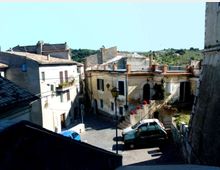 Veduta dall’alto di una piccola piazza in un borgo con case in muratura intonacata e tetti in tegole, balconi e finestre con persiane. Una strada in pendenza con lampione e auto parcheggiate attraversa lo spazio, con colline verdi sullo sfondo.