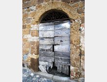 Porta ad arco in legno molto usurato e parzialmente rotto, inserita in una muratura in pietra con cornice in blocchi e piccoli inserti in mattoni. Sopra la porta c’è una grata in ferro battuto decorativa, con pavimentazione in pietra e ciottoli davanti all’ingresso.