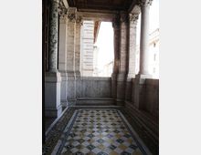 Loggia in pietra di un edificio storico con colonne e capitelli decorati, soffitto a cassettoni e parapetto alto. Il pavimento è rivestito da un mosaico geometrico a rombi con bordura ornamentale, affacciato sulla strada e su altri edifici.