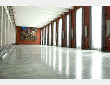 Ampia sala interna con pavimento in marmo lucido e soffitto a travi bianche, affacciata su una lunga fila di alte finestre con tende grigie. Pareti color terracotta con applique e, in fondo, un grande dipinto astratto.