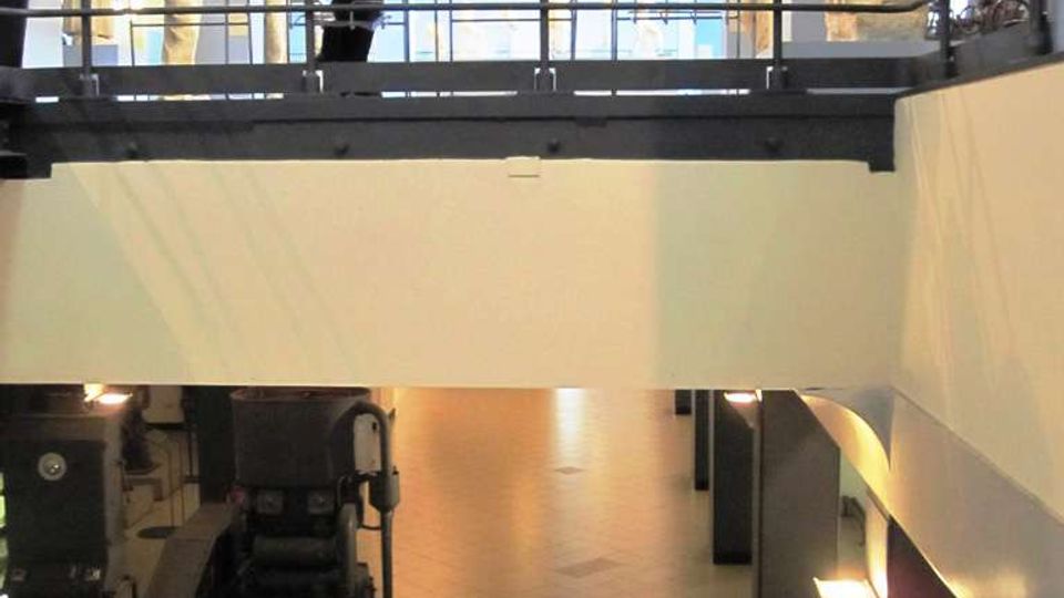 Interno di un museo industriale su due livelli, con soppalco in metallo e ringhiere sopra una grande sala. Al piano terra si vedono macchinari storici e una scala metallica, mentre in alto ci sono sedute e una statua vicino a grandi finestre e travi a vista.