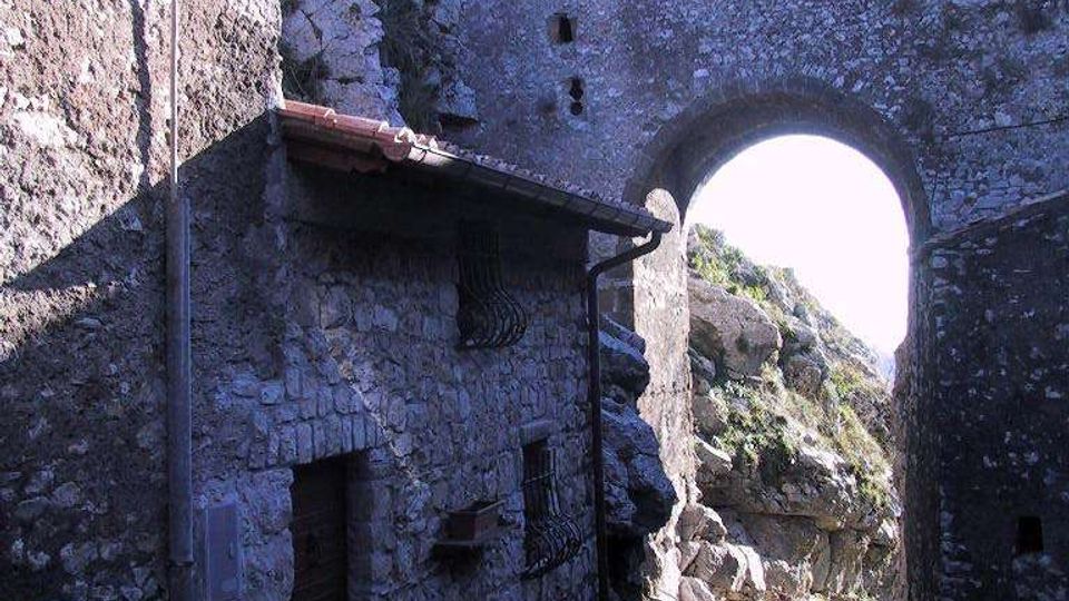 Vicolo stretto in pietra con pavimentazione acciottolata, fiancheggiato da edifici in muratura grezza con piccole finestre e una lanterna a parete. In fondo si apre un grande arco in pietra, che incornicia rocce e vegetazione su un pendio esterno.
