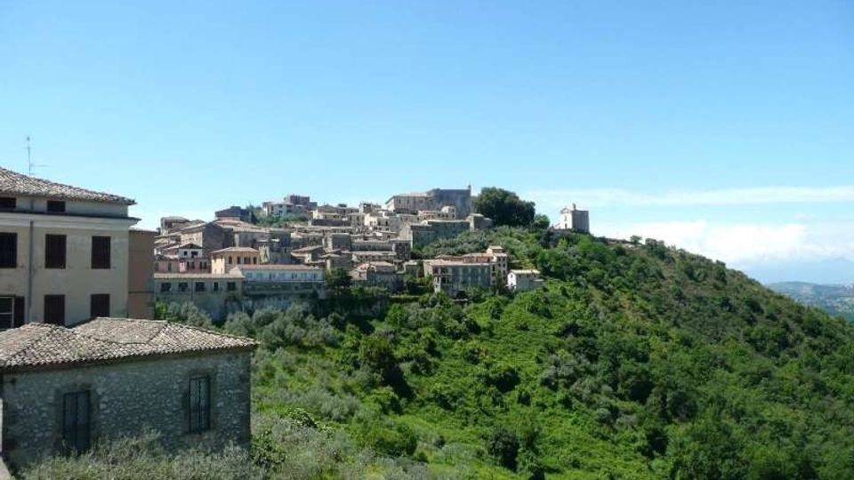 Veduta di un borgo collinare con case in pietra e tetti in coppi raggruppate lungo la cresta. In primo piano si vedono edifici con muratura in pietra, mentre intorno si estende un pendio verde ricoperto di vegetazione sotto un cielo sereno.