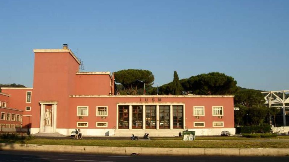 Edificio basso in stile razionalista con facciata rosa, ingresso centrale con grandi vetrate e gradinata, affiancato da un volume più alto sul lato sinistro. Davanti c’è una strada ampia con prato e alcuni veicoli, sullo sfondo alberi e un ponte metallico sulla destra.