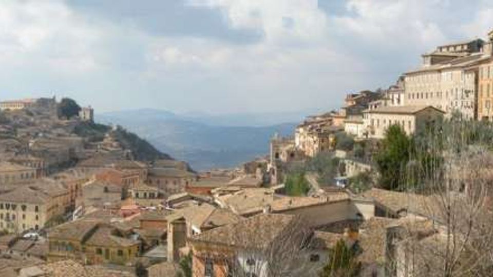 Panorama di un borgo collinare con case in pietra dai tetti in tegole, addossate lungo un crinale e su pendii terrazzati. Sullo sfondo si vedono colline e vallate, con rami spogli in primo piano e un gruppo di edifici più grandi in cima al colle.