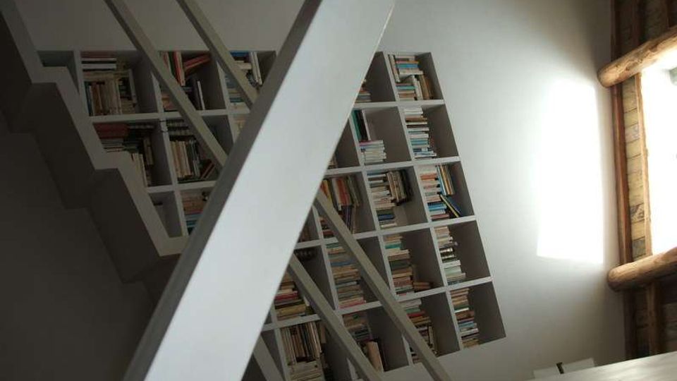 Interno di una stanza con una grande libreria a griglia piena di libri, vista in diagonale dietro una struttura metallica di scale o soppalco. A destra, una finestra con cornice in legno lascia entrare luce naturale sulla parete chiara.