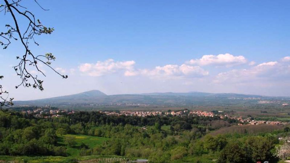 Veduta panoramica di una valle verde con campi e boschi, punteggiata da piccoli centri abitati e strade. Sullo sfondo si vedono colline e montagne basse sotto un cielo azzurro con alcune nuvole.