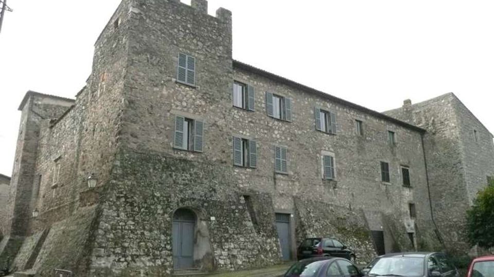 Edificio storico in pietra simile a un castello, con torre angolare merlata, mura inclinate e finestre con persiane. Davanti si vede un piccolo parcheggio con auto e una strada in pavé ai piedi della struttura.
