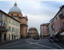 Piazza lastricata in pietra con fontana centrale, circondata da edifici storici a più piani con persiane e balconi. Sul lato sinistro domina una grande chiesa con facciata in pietra e ampia cupola.