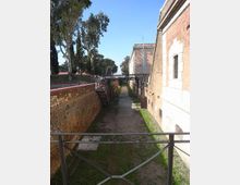 Fossato asciutto di una fortificazione, con percorso in terra e tratti d’erba tra alte mura in mattoni e pietra. In fondo si vede un ponte pedonale in ferro; sul lato sinistro ci sono alberi e una scalinata che scende nel fossato.