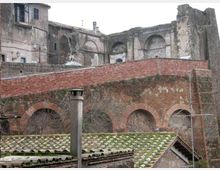 Rovine di un complesso storico in muratura con grandi arcate e nicchie, affiancate da edifici in pietra. In primo piano si vede un muro di contenimento in mattoni sopra una struttura ad archi e tetti con tegole verdi.
