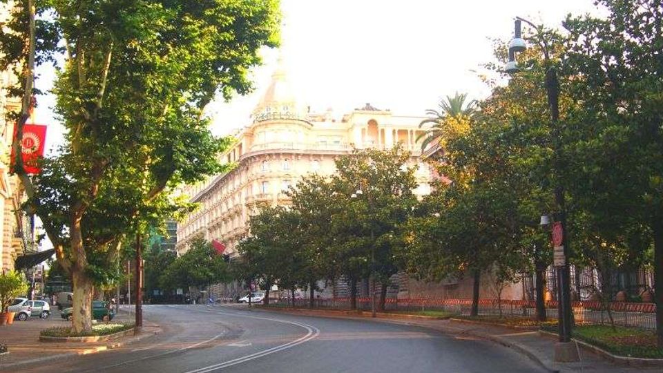 Viale urbano ampio e curvo, fiancheggiato da alberi e lampioni, con una recinzione lungo il marciapiede. Sullo sfondo si vede un grande edificio storico chiaro con facciata decorata e cupola.