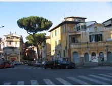 Incrocio urbano con edifici storici a più piani dalle facciate intonacate e finestre con persiane, con archi al piano terra e antenne sui tetti. Strada ampia con auto parcheggiate, strisce pedonali in primo piano e un grande pino domestico che svetta tra i palazzi.