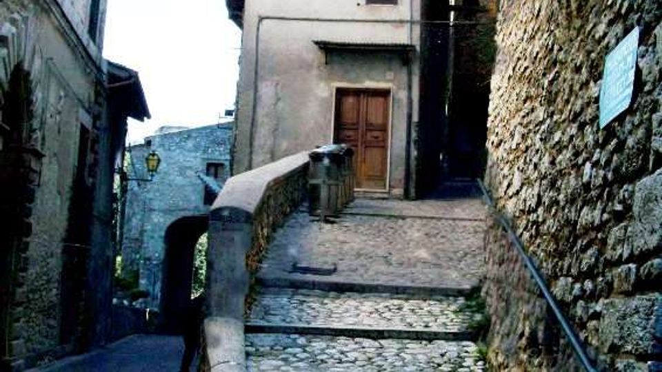 Vicolo stretto di un borgo storico con scalinata in ciottoli e muretto curvo, affiancata da edifici in pietra e intonaco con persiane in legno. Lampioni a parete e una piccola rampa sopraelevata conducono verso una casa con porta in legno.
