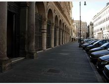 Portico urbano con lunga fila di arcate in pietra e colonne su un ampio marciapiede lastricato, affiancato da una strada con auto parcheggiate e lampioni. Sullo sfondo si vedono edifici storici e una piazza all’incrocio.