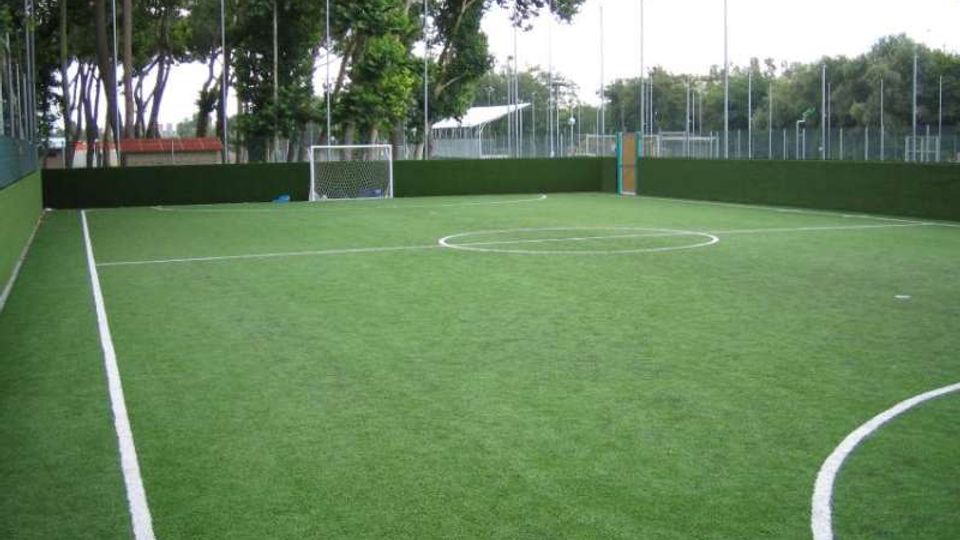 Campo da calcetto in erba sintetica con linee bianche e piccola porta sul fondo, circondato da recinzioni e pannelli verdi. Sullo sfondo si vedono alberi alti e pali dell’illuminazione lungo il perimetro.