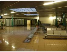 Ampia sala d’attesa interna con pavimento piastrellato lucido, file di sedute metalliche e colonne verdi. Il soffitto è illuminato da luci lineari e pannelli luminosi, con pareti vetrate e alcune piante in vaso lungo i lati.