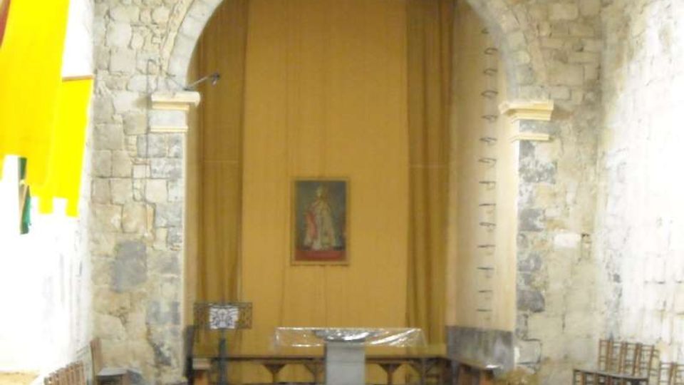 Interno di una piccola chiesa in pietra con grande arco centrale e pareti in muratura a vista. In fondo si vedono l’altare semplice, una pedana e sedie in legno disposte lungo i lati, con un dipinto appeso sulla parete posteriore.