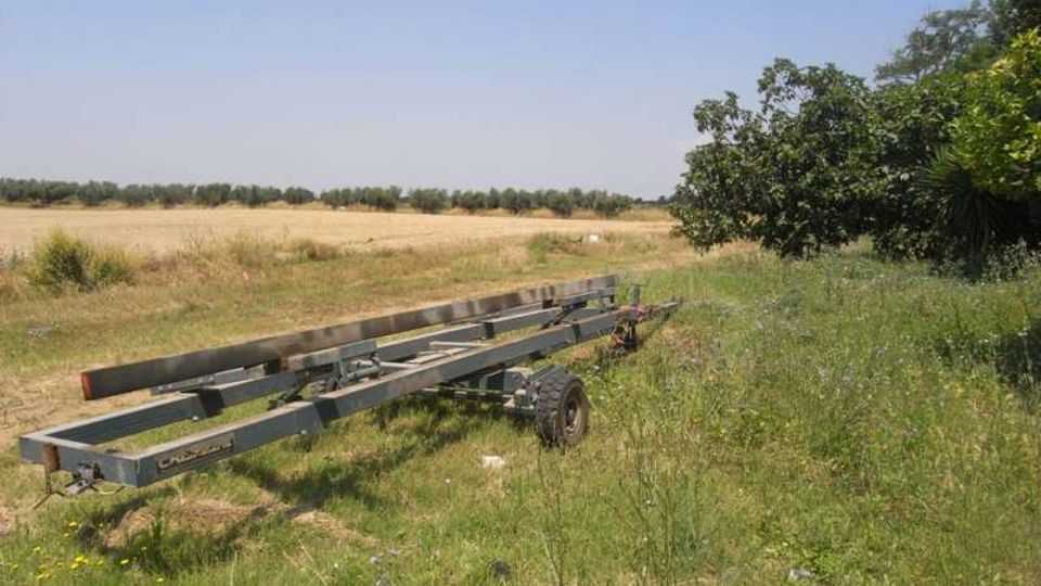 Campo rurale con erba alta e una lunga struttura metallica su ruote appoggiata a terra in primo piano. Sullo sfondo si vedono distese di campi coltivati e una linea di alberi sotto un cielo sereno.