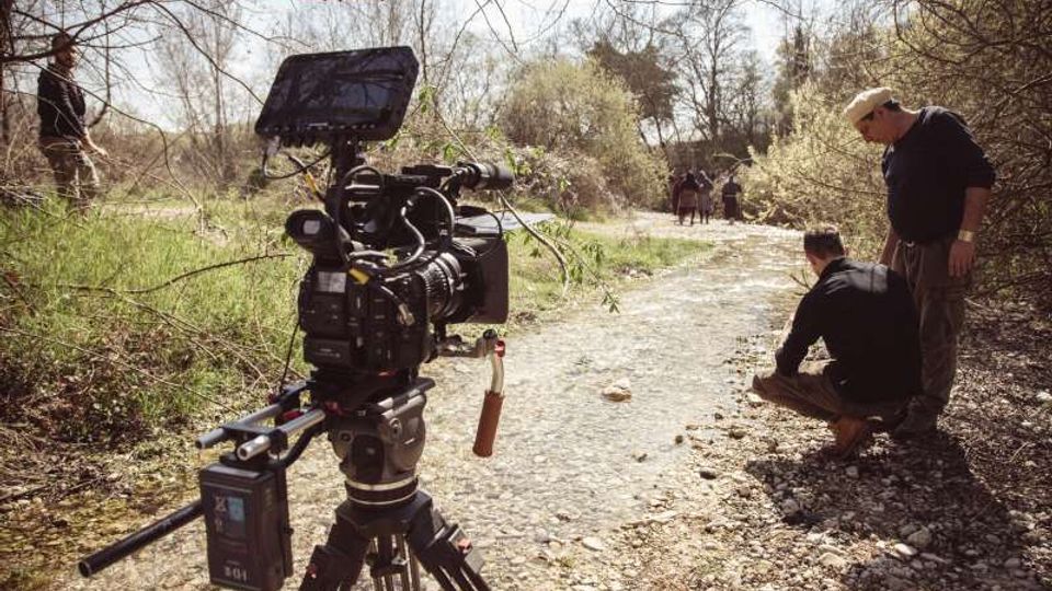 Sentiero sterrato e ghiaioso in un’area boschiva con vegetazione rada e alberi spogli, attraversato da un piccolo corso d’acqua poco profondo. In primo piano è presente una videocamera professionale su treppiede, con attrezzatura di ripresa lungo il bordo del sentiero.