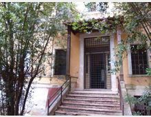 Ingresso di un edificio in muratura con intonaco color ocra, raggiunto da una scalinata con corrimano e coperto da una piccola tettoia. La porta è protetta da una grata metallica e la facciata è parzialmente nascosta da alberi e vegetazione fitta.