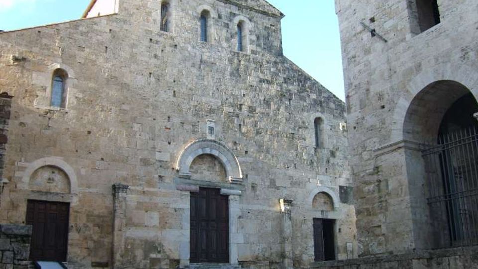 Facciata di una chiesa romanica in pietra con portale ad arco e piccole finestre strette, affacciata su una piazza con gradini in pietra. A destra si vede una struttura adiacente con grandi arcate e una cancellata in ferro.