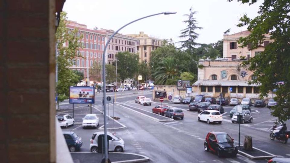 Ampio incrocio urbano con più corsie e parcheggi, con auto e strisce pedonali visibili. Sullo sfondo si vedono edifici multipiano, un edificio basso con finestre ad arco e murales, e alberi tra cui una palma e una conifera.