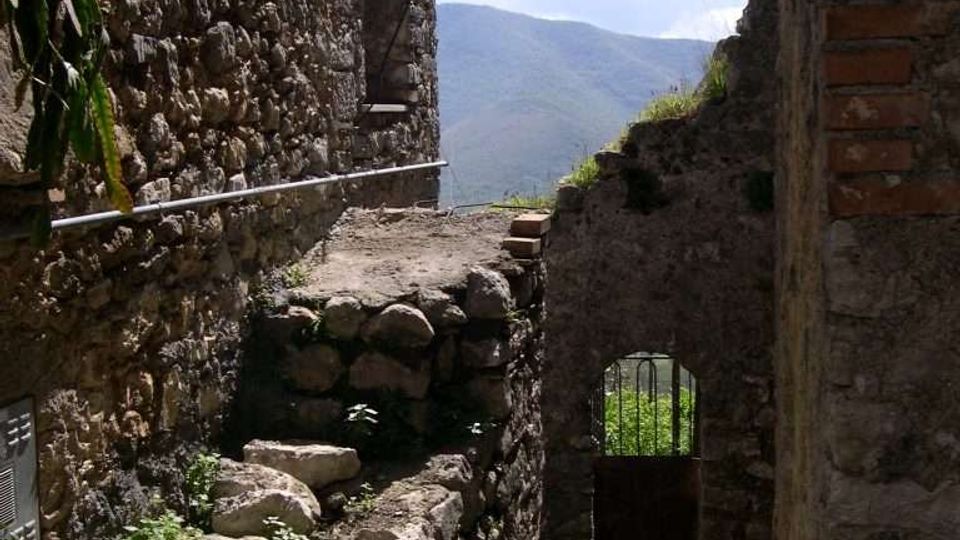 Stretto vicolo in pietra tra edifici rustici, con muri in pietra a vista e piccole piante lungo il percorso. In fondo si vede un passaggio ad arco con cancello e, oltre, una vista sulle montagne sotto un cielo sereno.