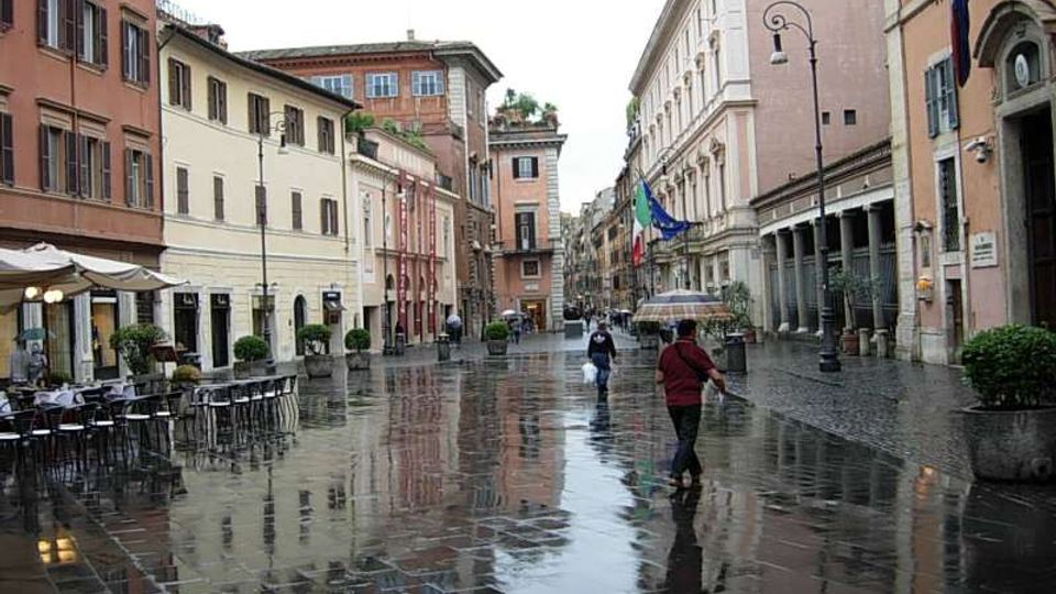 Ampia piazza urbana con pavimentazione in pietra bagnata dalla pioggia, circondata da edifici storici dai colori pastello e facciate con finestre regolari. Ai lati si vedono tavolini di un bar con ombrelloni, portici con colonne, lampioni e grandi fioriere.