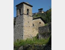Campanile in pietra con aperture ad arco e campana visibile, accanto a una piccola chiesa con facciata intonacata e oculo. L’edificio sorge su un pendio erboso con muri in pietra e vegetazione, con case e alberi sullo sfondo sotto un cielo sereno.