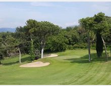Campo da golf con fairway in erba rasata, green e due bunker di sabbia, circondato da alberi alti e vegetazione fitta. Sullo sfondo si intravedono colline sotto un cielo sereno.