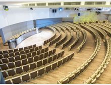 Interno di un’aula magna moderna con pavimento in legno, file di sedute disposte a semicerchio e una postazione relatori con tavolo e podio. Pareti chiare con pannelli acustici, illuminazione da soffitto e balconata laterale sopraelevata.